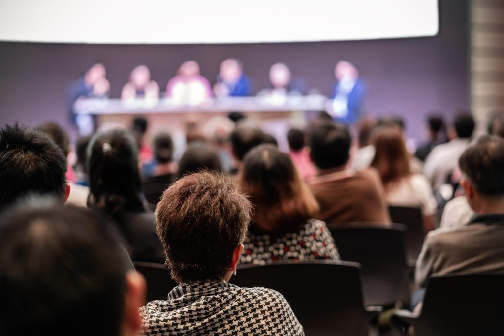 Rear view of audience in a conference hall where speakers are talking on the stage.